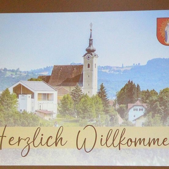 Eine Kirche mit Turm befindet sich im Dorf, umgeben von Bäumen und Bergen. Der Text 'Herzlich Willkommen' ist auf Deutsch geschrieben.
