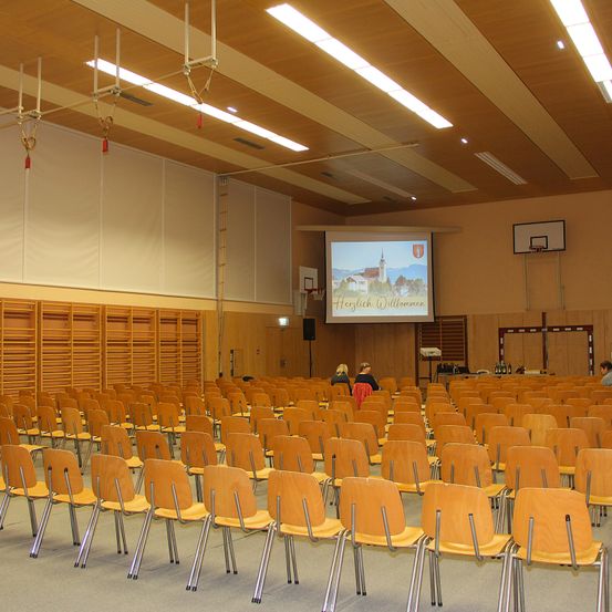Ein leerer Konferenzraum mit Holzstühlen, einer Projektionsleinwand und zwei Personen, die in der Ecke sitzen. Der Raum hat einen Basketballkorb, einen Lautsprecher und einen Projektor.
