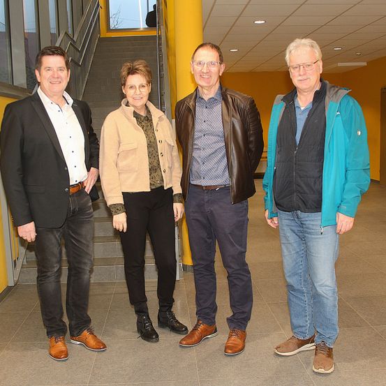 Vier Personen stehen in einem Flur zusammen und tragen Freizeitkleidung. Sie lächeln und scheinen für ein Foto zu posieren. Hinter ihnen befindet sich eine Treppe und eine gelbe Säule.