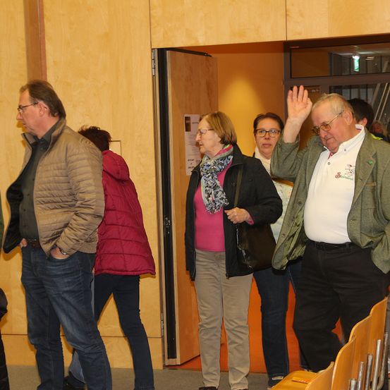 Eine Gruppe von Menschen in einem Raum. Ein älterer Mann winkt mit der Hand. Eine Frau mit einer roten Jacke und eine Frau mit Brille und Schal sind anwesend.