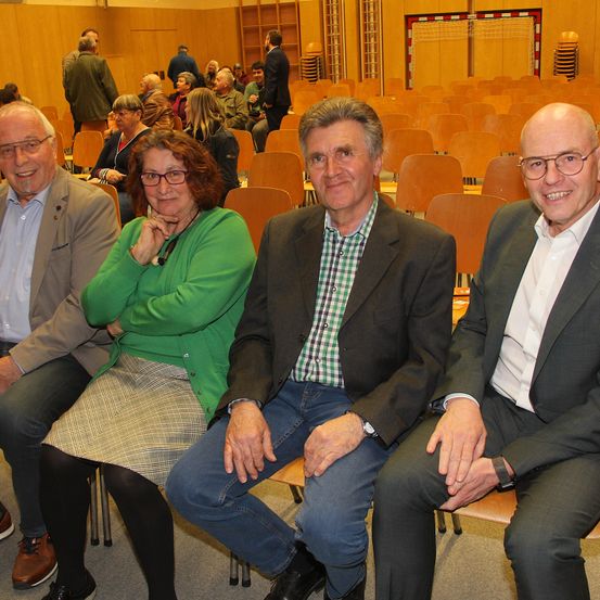 Vier Erwachsene, zwei Männer und zwei Frauen, sitzen in einem Klassenzimmer. Sie scheinen für ein Foto zu posieren. Hinter ihnen sitzen und stehen weitere Personen. Das Klassenzimmer hat Holzstühle, Wände und ein Tor im Hintergrund.