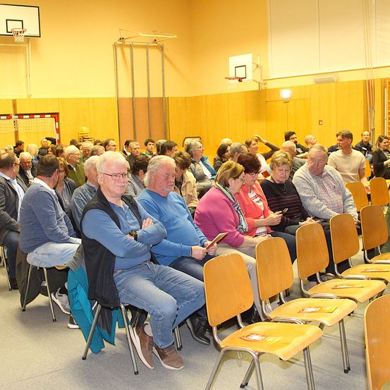 Eine Gruppe älterer Erwachsener, hauptsächlich Männer, sitzt in einer Turnhalle mit Holzwänden und Basketballkörben. Sie tragen Freizeitkleidung und manche tragen eine Brille. Stühle sind in Reihen aufgestellt, mit Broschüren darauf.