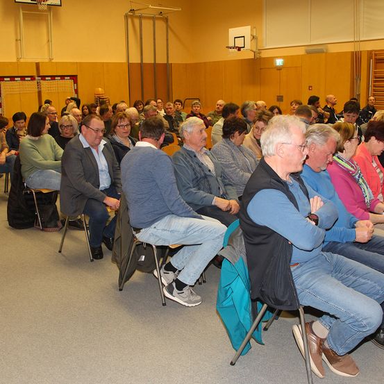 Ein Raum voller Menschen, meist ältere Erwachsene, die auf Stühlen sitzen. Sie scheinen an einer Versammlung oder einem Treffen teilzunehmen. Der Raum hat ein Basketballnetz und eine Holzwand.
