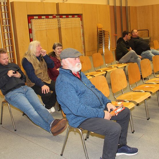 Eine Gruppe von Menschen sitzt in Stühlen in einem Raum. Ein Mann hält ein Handy, während ein anderer Mann etwas liest. Der Raum hat Holzwände und ein Tornetz.