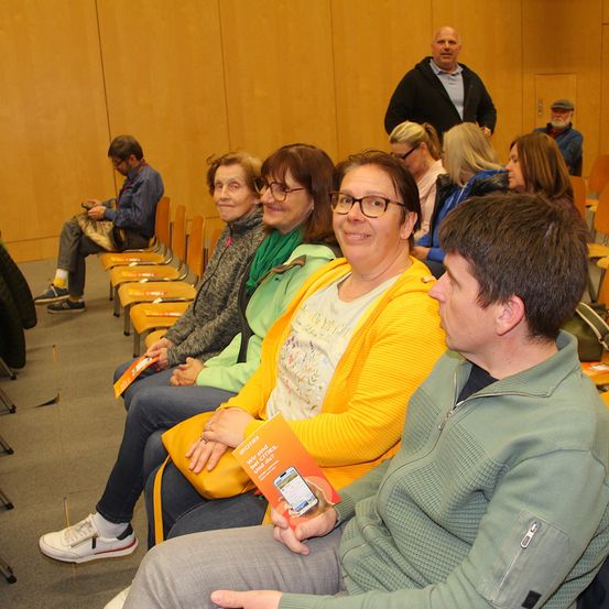 Eine Gruppe von Menschen sitzt in Stühlen in einem Besprechungsraum und schaut alle nach vorne. Ein Mann vorne schaut auf sein Handy. Einige Leute tragen Brillen. Hinter ihnen steht ein Mann.