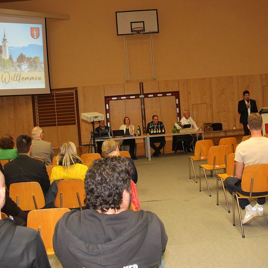 In einem Raum mit Holzwänden und Stühlen findet eine Konferenz statt. Eine Projektionsleinwand zeigt 'Willkommen'. Vier Personen sitzen an einem Tisch mit Flaschen, und ein Mann spricht in ein Mikrofon.
