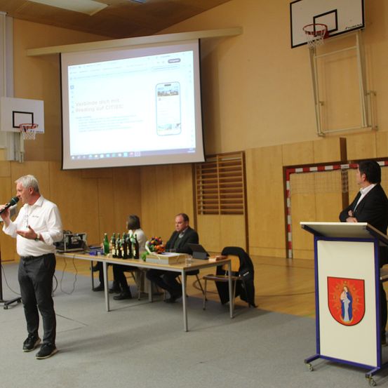 Ein Mann spricht in ein Mikrofon auf der Bühne. Eine Projektionsleinwand zeigt eine Folie. Zwei Personen sitzen an einem Tisch mit Flaschen und Blumen. Ein anderer Mann steht an einem Podest.