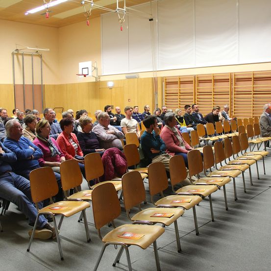 Eine Gruppe von Menschen sitzt in Reihen von Stühlen in einem großen, offenen Raum mit einem Basketballkorb. Sie sehen aufmerksam aus, möglicherweise warten sie darauf, dass eine Veranstaltung beginnt. Der Raum ist gut beleuchtet und hat Holzwände und -böden.
