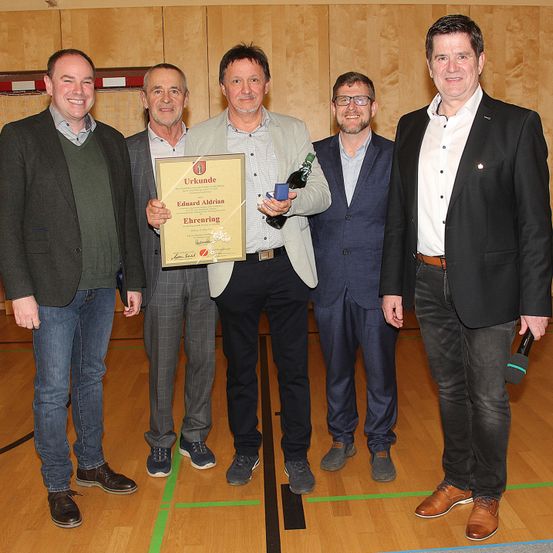 Fünf Männer stehen in einem Raum, einer hält Plakette und Flasche, lächeln. Dahinter eine Holzwand. Holzboden mit grünen Linien.