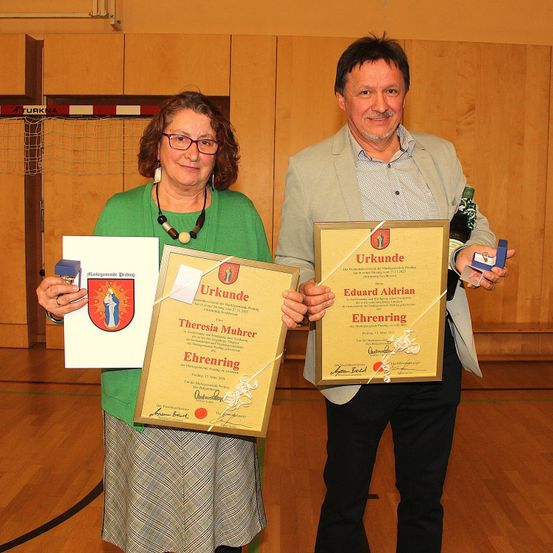 Zwei Personen, ein Mann und eine Frau, stehen und halten gerahmte Urkunden. Der Mann hält eine Flasche und eine kleine Trophäe. Sie lächeln beide und scheinen für ein Foto zu posieren. Hinter ihnen ist eine Holzwand mit einem Netz und einem darauf geschriebenen Wort.
