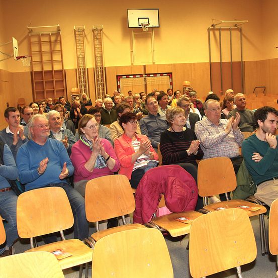 Eine Menschenmenge sitzt in Stühlen und klatscht in einer Innenhalle mit Holzwänden und Basketballausrüstung.