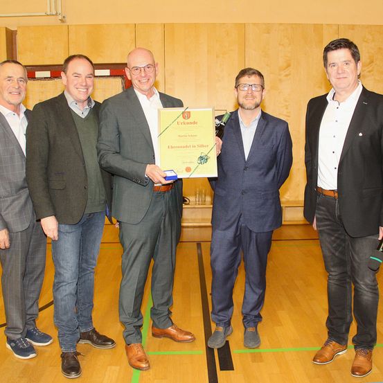 Fünf Männer stehen in einem Raum zusammen, einer hält ein gerahmtes Zertifikat und eine Flasche. Sie alle tragen formelle Kleidung, und es gibt einen Holzboden und Holzwände im Hintergrund.