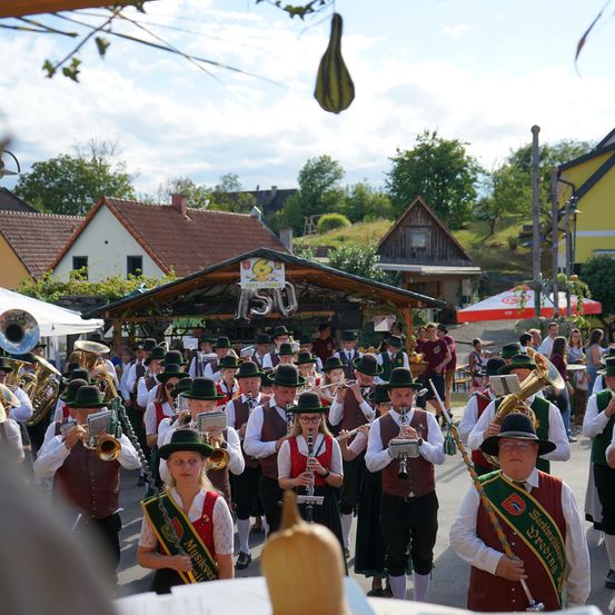 Eine große Gruppe von Musikern in traditioneller bayerischer Kleidung, einschließlich grüner Hüte und Westen, tritt bei einem Freiluftumzug auf. Sie spielen verschiedene Instrumente wie Trompeten, Tuben und Flöten. Der Hintergrund zeigt ein sonniger Tag mit Häusern und Bäumen.