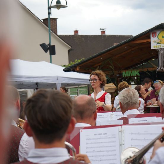 Eine Frau in einem roten Kleid steht vor einer Brassband und leitet sie während einer Aufführung auf einem Freiluftfestival.