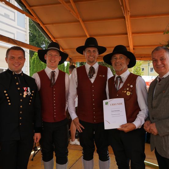 Fünf Männer in traditioneller Kleidung stehen unter einem Baldachin, einer hält ein Zertifikat. Sie lächeln und scheinen bei einer Veranstaltung zu sein.