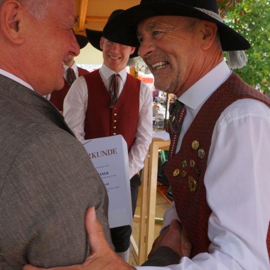 Zwei Männer in traditioneller Kleidung, einer hält ein Zertifikat, lächeln und schütteln sich die Hände im Freien.