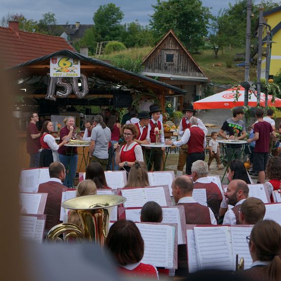 Eine Gruppe von Menschen in traditioneller Kleidung tritt in einer Freiluftveranstaltung auf, wobei Musiker Instrumente spielen und andere Essen an einem Stand servieren.