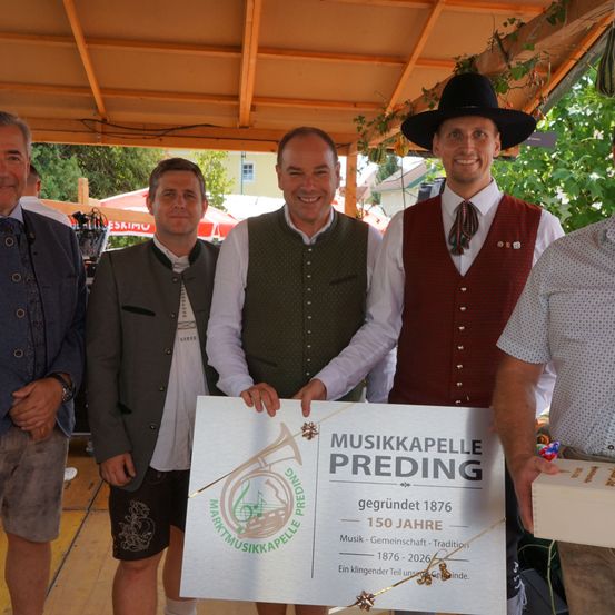 Fünf Männer in traditioneller deutscher Kleidung stehen unter einem Baldachin, lächeln und halten ein Zertifikat für die Musikkapelle Preding, die ihr 150-jähriges Jubiläum feiert.