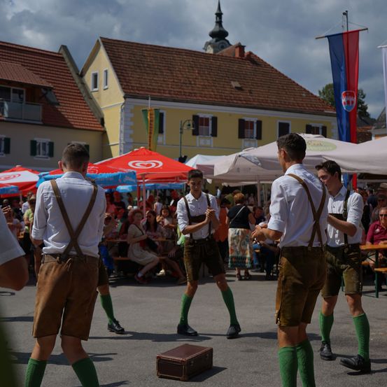 Vier Männer in traditioneller bayerischer Kleidung führen auf einer Straße vor einer Menge auf. Die Männer tragen Hosenträger und grüne Socken. Im Hintergrund befinden sich Zelte und Fahnen.