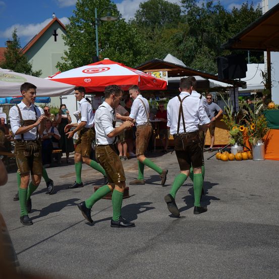 Eine Gruppe von Männern in traditioneller bayerischer Kleidung tanzt auf einem öffentlichen Platz. Sie tragen grüne Socken und braune Lederschuhe.