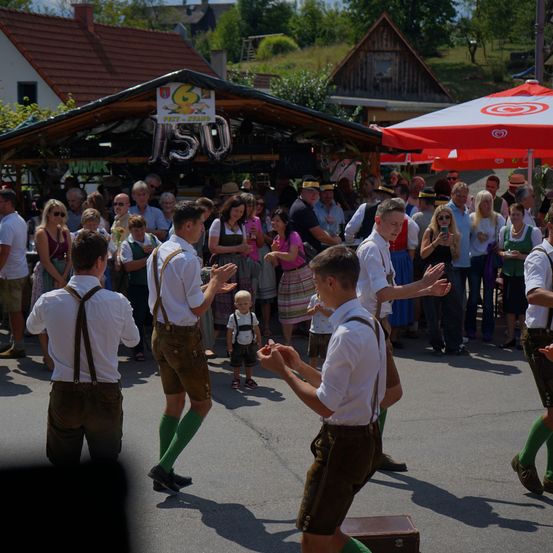 Eine Gruppe von Männern in traditioneller bayerischer Kleidung tanzt auf einem öffentlichen Platz, während eine Menge zuschaut. Ein rot-weißer Regenschirm ist im Vordergrund zu sehen.