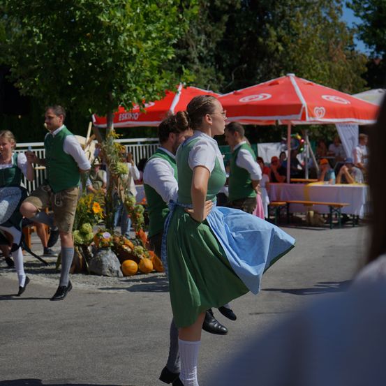 Eine Gruppe von Menschen in traditioneller bayerischer Kleidung tanzt im Freien, mit roten Sonnenschirmen und Kürbissen im Hintergrund.