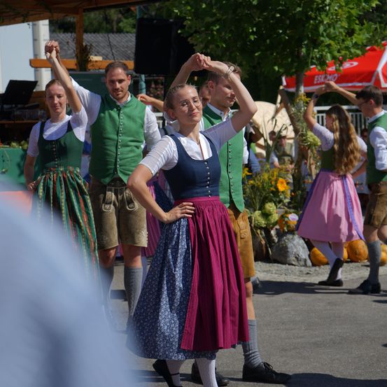 Eine Gruppe von Menschen in traditioneller bayerischer Kleidung tanzt im Freien. Die Frau in der Mitte trägt ein blaues Oberteil und einen braunen Rock. Hinter ihr halten ein Mann und eine Frau Händchen.