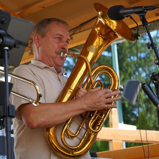 Ein Mann spielt eine goldene Tuba unter einem Holzvordach, mit Mikrofonen und einem Notenständer in der Nähe.