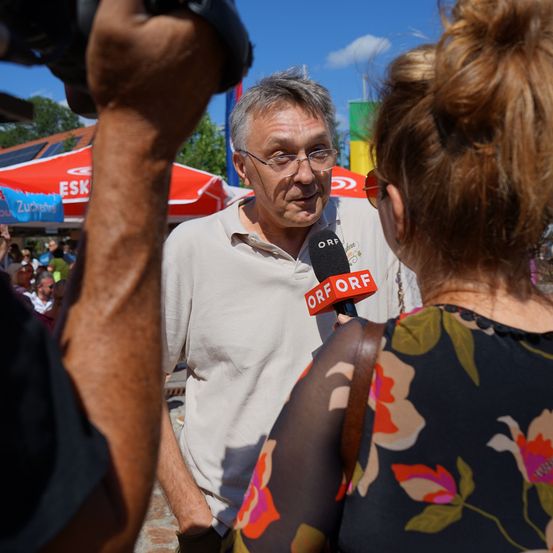 Ein Mann mit Brille wird von einer Frau mit einem Mikrofon bei einer öffentlichen Veranstaltung interviewt.
