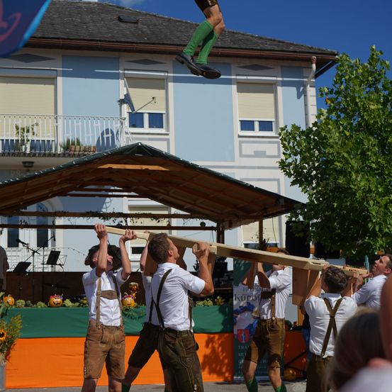 Eine Gruppe von Männern in traditioneller bayerischer Kleidung hebt eine lange Holzplanke vor einem Gebäude mit Bühne und Baum.