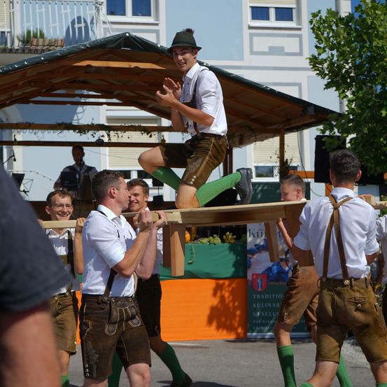 Eine Gruppe von Männern in traditioneller bayerischer Kleidung hebt eine Holzplanke, während ein Mann darauf balanciert. Sie treten bei einer Veranstaltung im Freien auf.