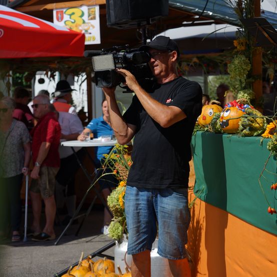 Ein Mann in einem schwarzen T-Shirt und blauen Shorts filmt mit einer Kamera bei einer Veranstaltung im Freien mit Kürbissen und Menschen im Hintergrund.