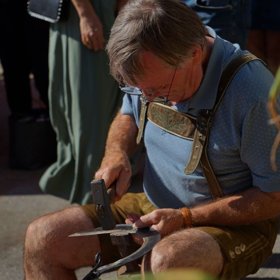 Ein älterer Mann in einem blauen Hemd und braunen Hosenträgern formt ein Metallwerkzeug mit einem Hammer.