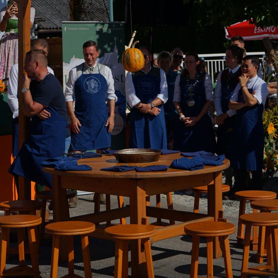 Eine Gruppe von Menschen in blauen Schürzen versammelt sich um einen Holztisch mit einer Schüssel und einer Kürbis, der darüber hängt. Sie nehmen wahrscheinlich an einem Kochwettbewerb teil.
