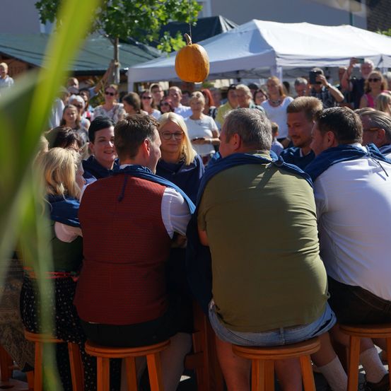 Eine Gruppe von Menschen sitzt auf Hockern in einer Außenumgebung, einige in traditioneller Kleidung. Ein Kürbis hängt von einem Zelt, und eine Menschenmenge versammelt sich unter dem Zelt.