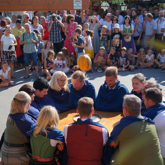 Eine Gruppe von Menschen, die in blaue und grüne Schals gekleidet sind, versammelt sich um einen Tisch, während eine Menge von Zuschauern hinter ihnen steht und zusieht. Einige der Zuschauer halten Kameras und machen Fotos.