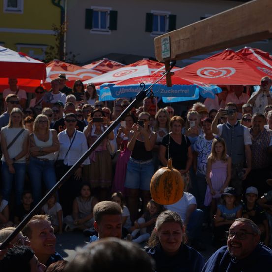 Eine Menschenmenge, die sich um eine an einem Seil hängende Kürbis versammelt hat, wahrscheinlich auf einem Festival. Einige machen Fotos mit ihren Handys.