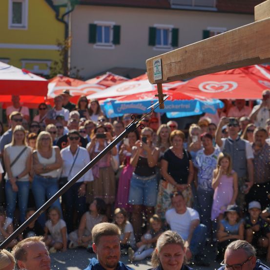 Eine Menschenmenge versammelt sich um eine Holzstruktur, möglicherweise für ein Bieranzapfen-Event, mit Schirmen und Gebäuden im Hintergrund.