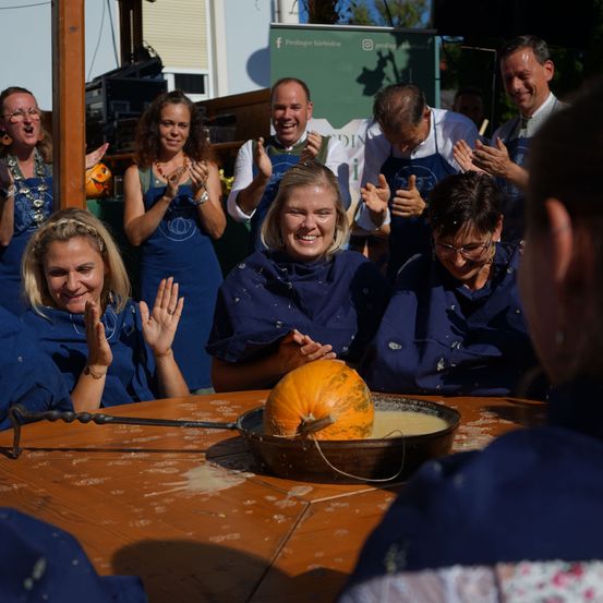 Eine Gruppe von Menschen in blauen Schürzen versammelt sich um einen Kürbis auf einem Tisch, jubelt und klatscht. Der Kürbis wird über einem Feuer geröstet.