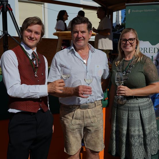 Drei Menschen stehen zusammen, lächeln und halten Weingläser. Der Mann in der Mitte trägt ein weißes Hemd und Shorts, während die anderen traditionelle Kleidung tragen.