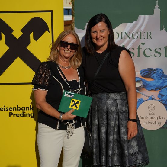 Zwei Frauen stehen vor einem Banner mit gelbem Hintergrund und schwarzem X-Symbol. Die Frau auf der linken Seite hält eine grüne Mappe mit einem gelben X darauf.