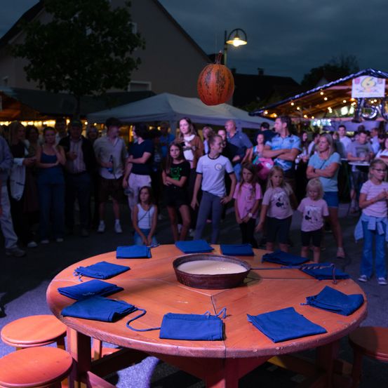 Eine Gruppe von Menschen versammelt sich um einen Tisch mit einer Schüssel Essen in der Mitte, mit einem Kürbis darüber. Sie stehen in einem Außenbereich bei Nacht.