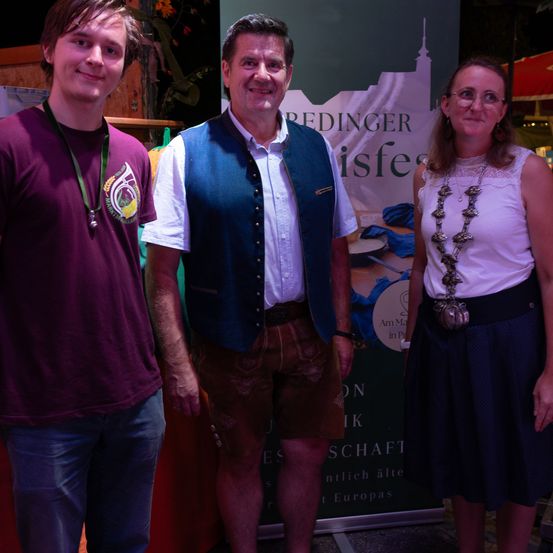 Drei Personen stehen gemeinsam vor einem Banner, zwei Männer und eine Frau, alle lächeln für die Kamera. Der Mann auf der linken Seite trägt ein burgunderrotes Shirt und einen grünen Ausweis. Der Mann in der Mitte trägt eine blaue Weste über einem weißen Hemd und braunen Shorts. Die Frau auf der rechten Seite trägt ein weißes Oberteil, eine Halskette und einen blauen Rock.