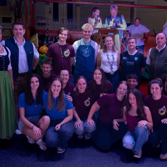 Eine Gruppe von Menschen in Freizeitkleidung, einige mit Schürzen, posiert für ein Foto in einer festlichen Umgebung mit einer Bühne und Musikinstrumenten im Hintergrund.
