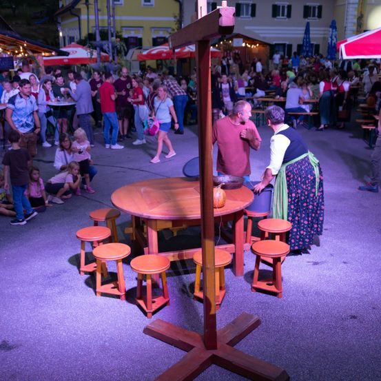 Eine Gruppe von Menschen versammelt sich um einen Holztisch mit einem Kürbis darauf, in einer Außenumgebung. Der Tisch ist von roten Hockern umgeben. Im Hintergrund sitzen und stehen viele Menschen, einige unter Schirmen.