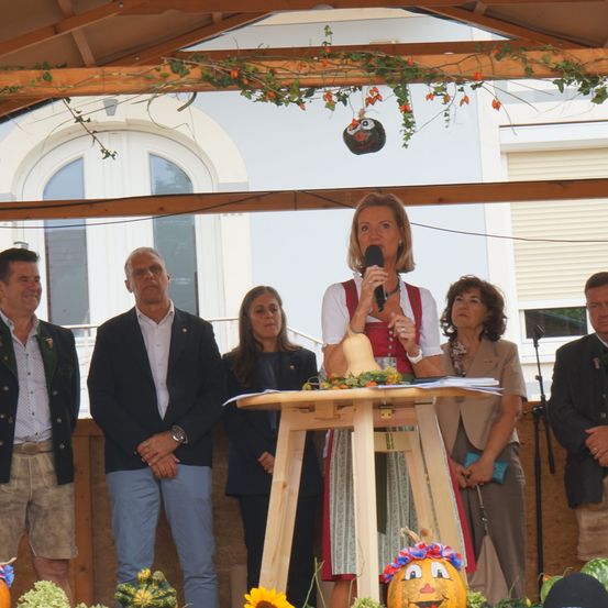 Eine Frau spricht in ein Mikrofon an einem Podium, umgeben von Menschen in traditioneller Kleidung. Hinter ihr ist ein Gebäude mit Bogenfenstern und einem dekorativen Kürbis zu sehen.