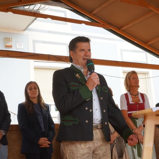 Ein Mann in traditioneller bayerischer Kleidung spricht in ein Mikrofon bei einer Veranstaltung im Freien, mit zwei Frauen hinter ihm.