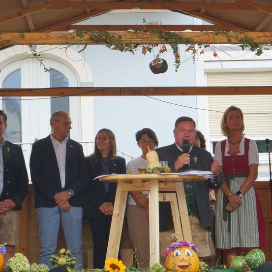 Ein Mann spricht in ein Mikrofon auf einem Podium mit Kürbis und Gourden, umgeben von Menschen in traditioneller deutscher Kleidung.