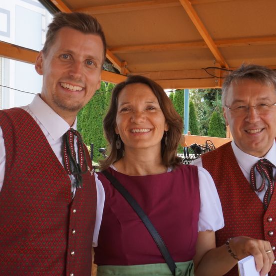 Drei Personen in traditioneller Kleidung, die lächelnd für ein Foto im Freien posieren.