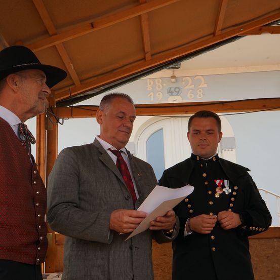 Drei Männer in formeller Kleidung stehen unter einem hölzernen Vordach. Der Mann in der Mitte hält ein Stück Papier, während der Mann rechts eine Militäruniform mit Medaillen trägt.